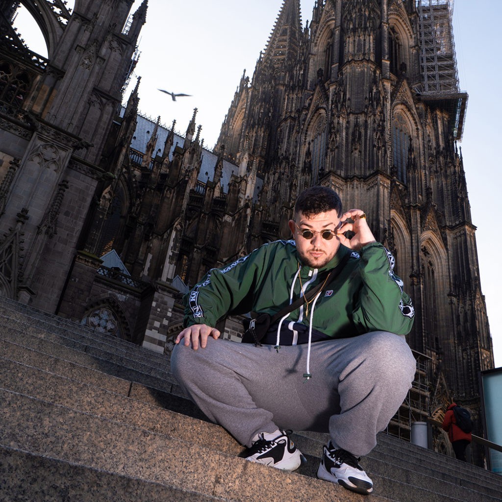 KABU sitz auf den Stufen vor dem Kölner Dom und schaut in die Kamara. Er hat eine Sonnenbrille auf und eine grüne Jacke an.