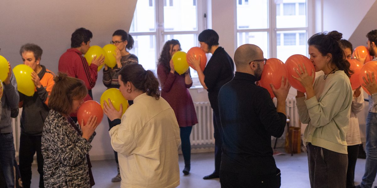 Many people stand together in pairs holding balloons to their lips.